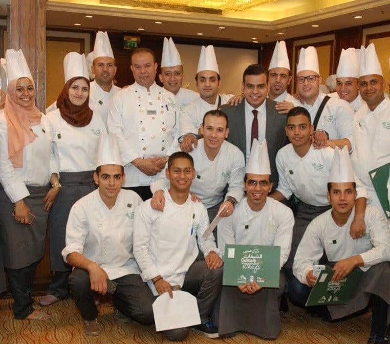 A graduating group of chefs in whites and toques, standing with Alaa after a ceremony