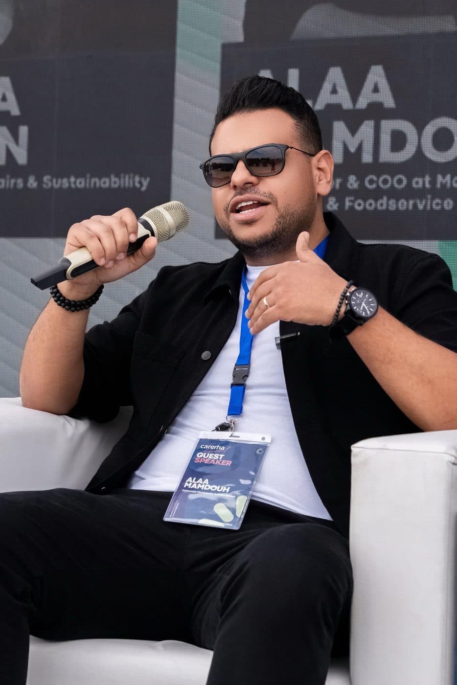 Alaa speaking with a microphone on stage, his name on the backdrop behind him